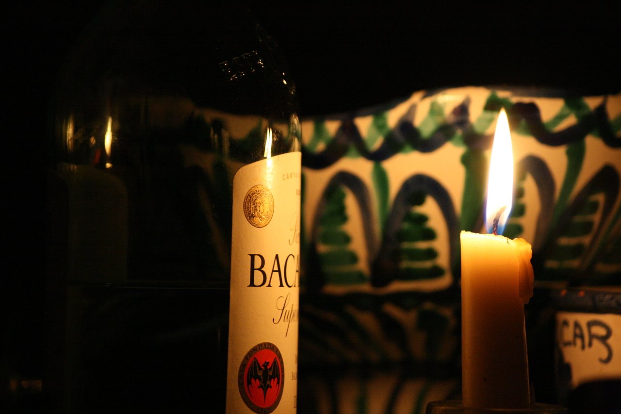 Photo description: Imagen oscura con una botella de ron, una vela encendida y dos piezas de cerámica granadina: una de azúcar y un frutero sin fruta.