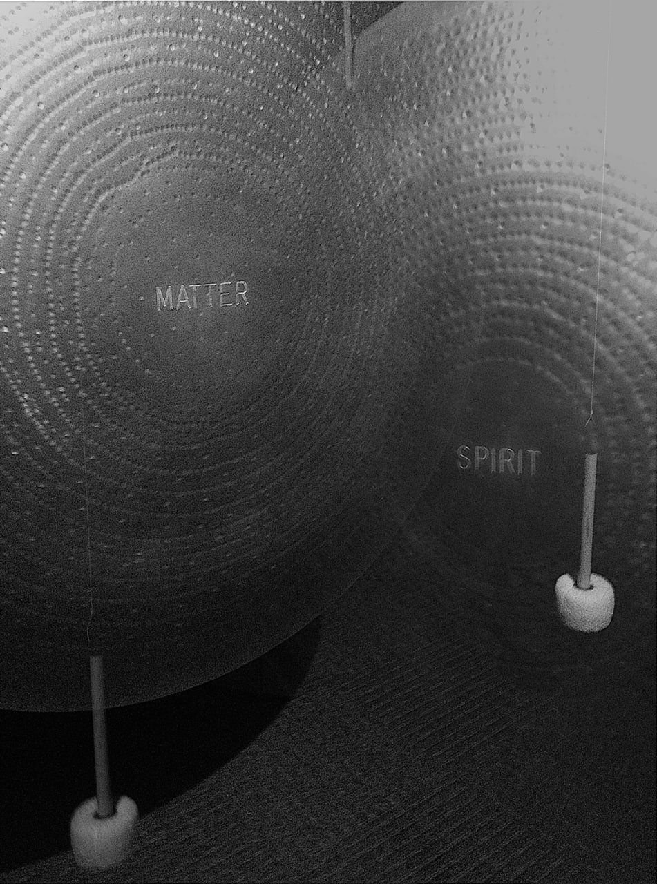 Photo description: Superposition of two images, each depicting a large bronze tam-tam (gong). They have a word engraved on their centres, 'matter' in one and 'spirit' in the other. Each one has its own mallet hanging from a thread.